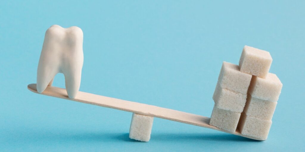 A seesaw made of a wooden stick balanced on a sugar cube, with a large white tooth on one side and stacked sugar cubes on the other, symbolizing dental myths that people shouldn’t believe, such as sugar being the sole cause of cavities.